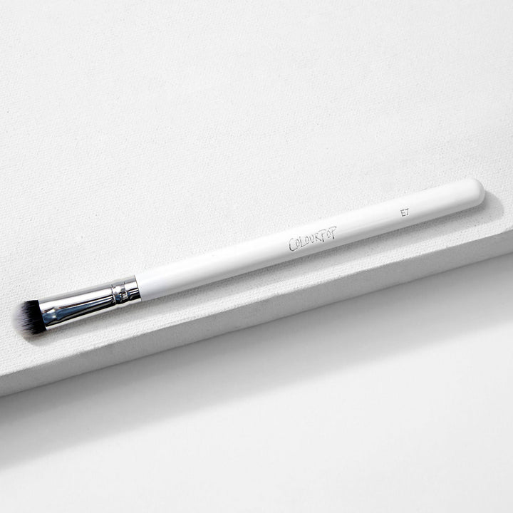 ColourPop Medium Dome Brush blending detail eye makeup - white-handled makeup brush with chrome ferrule and domed black‑to‑white bristles, marked E7 - resting diagonally on a minimalist white textured backdrop.