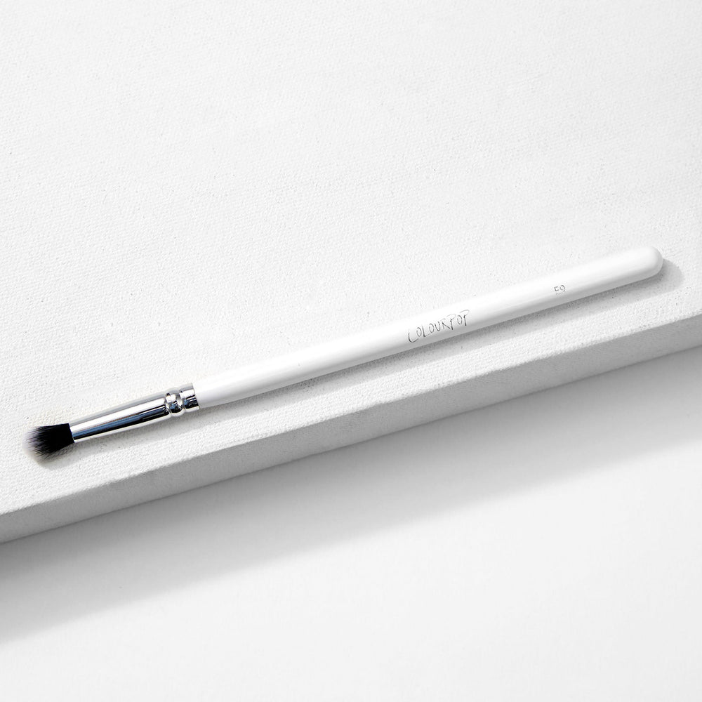 ColourPop Small Tapered Brush resting diagonally on a white textured platform, featuring a slim white handle, chrome ferrule, and small tapered black-and-white bristles for precise eyeshadow blending and detail work.