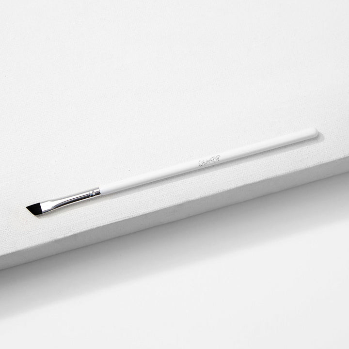 ColourPop Angled Eye Brush resting diagonally on a white textured block, featuring slim angled black bristles, a silver ferrule, and a long white handle - makeup brush for precise eyeliner and brow shaping.