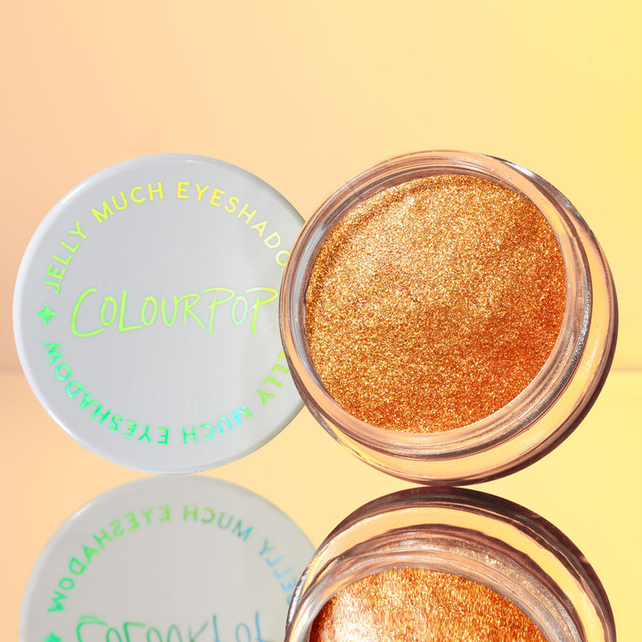 ColourPop Jelly Much Shadow in Brentwood sits open, showcasing a metallic copper-gold jelly eyeshadow with microglitter; white lid with neon green logo beside it, reflected on a glossy surface against a warm peach backdrop.