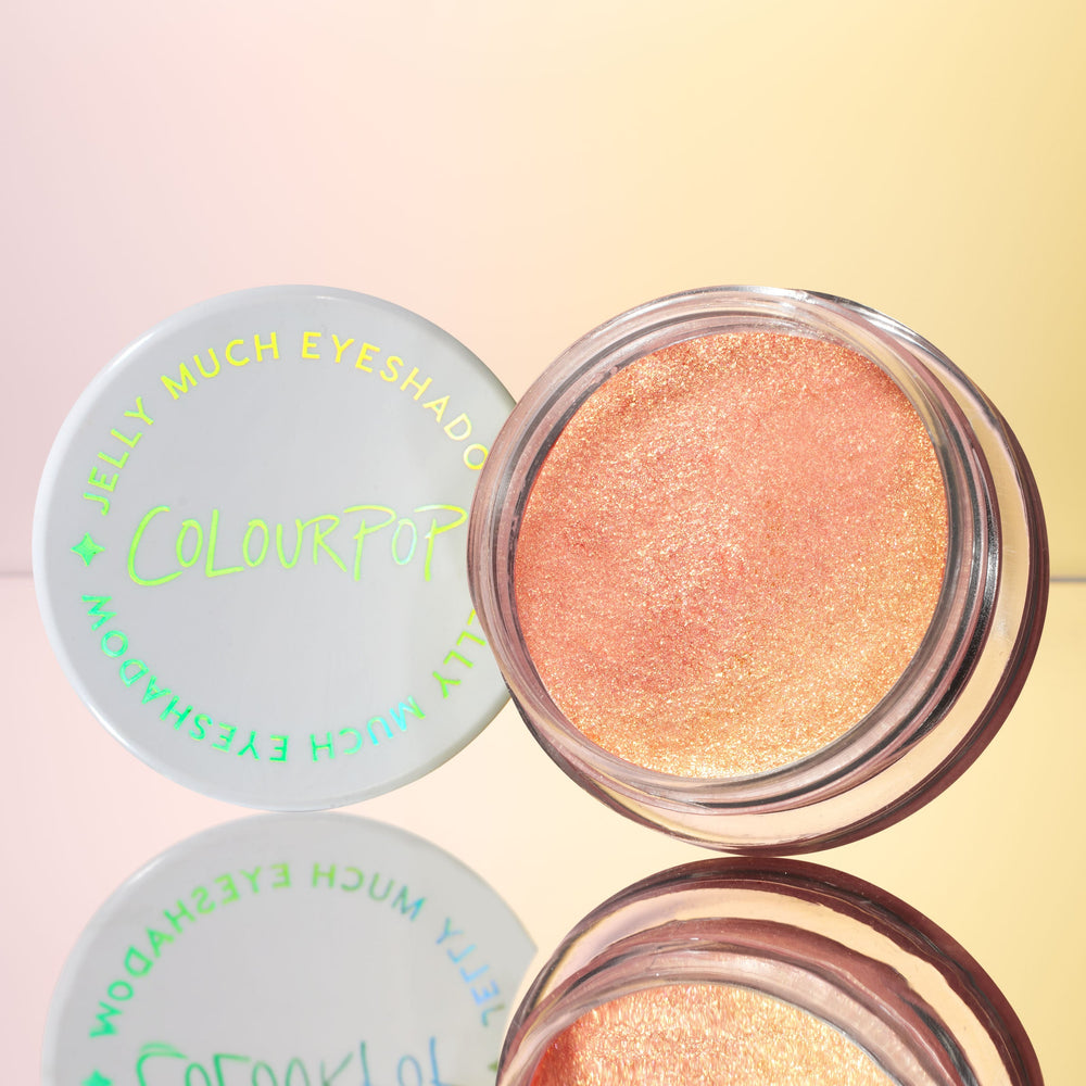 Open pot of ColourPop Jelly Much Shadow in Buzzy, a peachy coral-gold shimmer, sitting beside a white lid with iridescent lettering on a mirrored surface against a warm gradient backdrop.