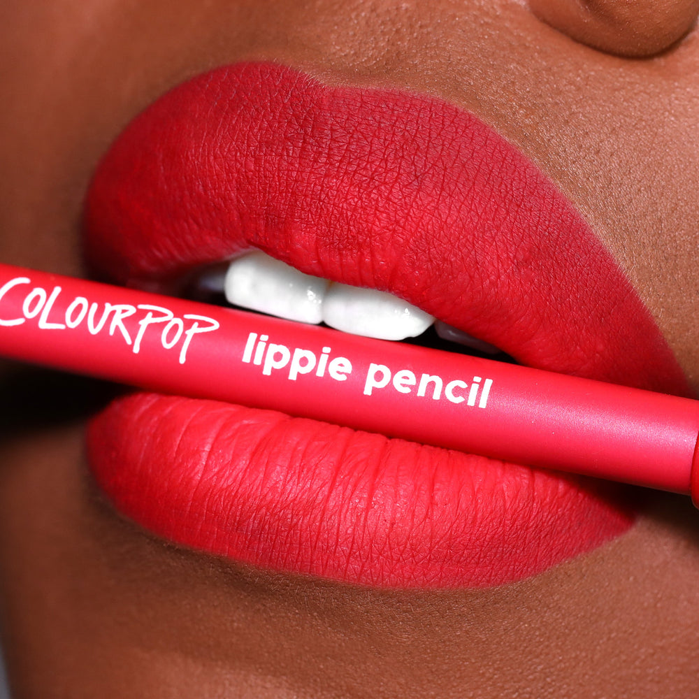 ColourPop Lippie Pencil in Overtime held between vividly lined, matte red lips, showcasing the creamy lip liner’s precise definition in a close-up beauty shot with the hot-pink barrel and white logo text visible.