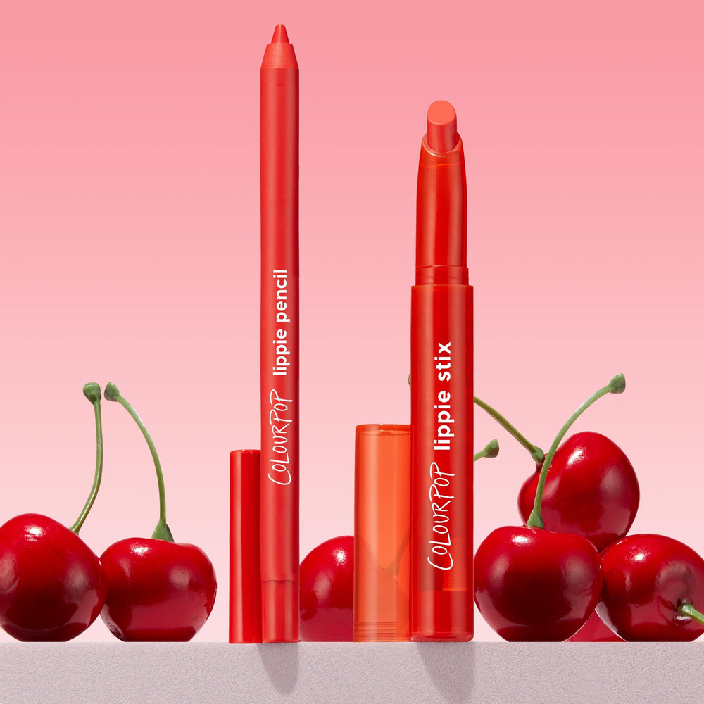ColourPop Chi Chi Lippie Stix and ColourPop Lippie Pencil stand uncapped to show vivid red tips, set among glossy cherries on a pink gradient background, highlighting the bright cherry-red lip duo for bold, defined color.