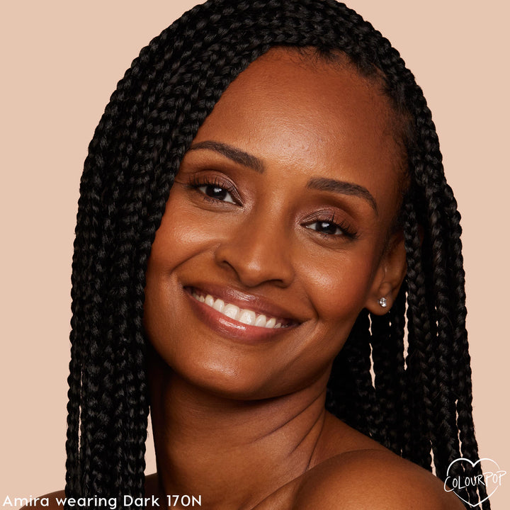 Model wearing ColourPop Pretty Fresh Creamy Concealer, shade Dark 170 N, smiles to showcase smooth, even under-eye coverage and a dewy, natural finish; close-up with braided hair against a warm beige backdrop.