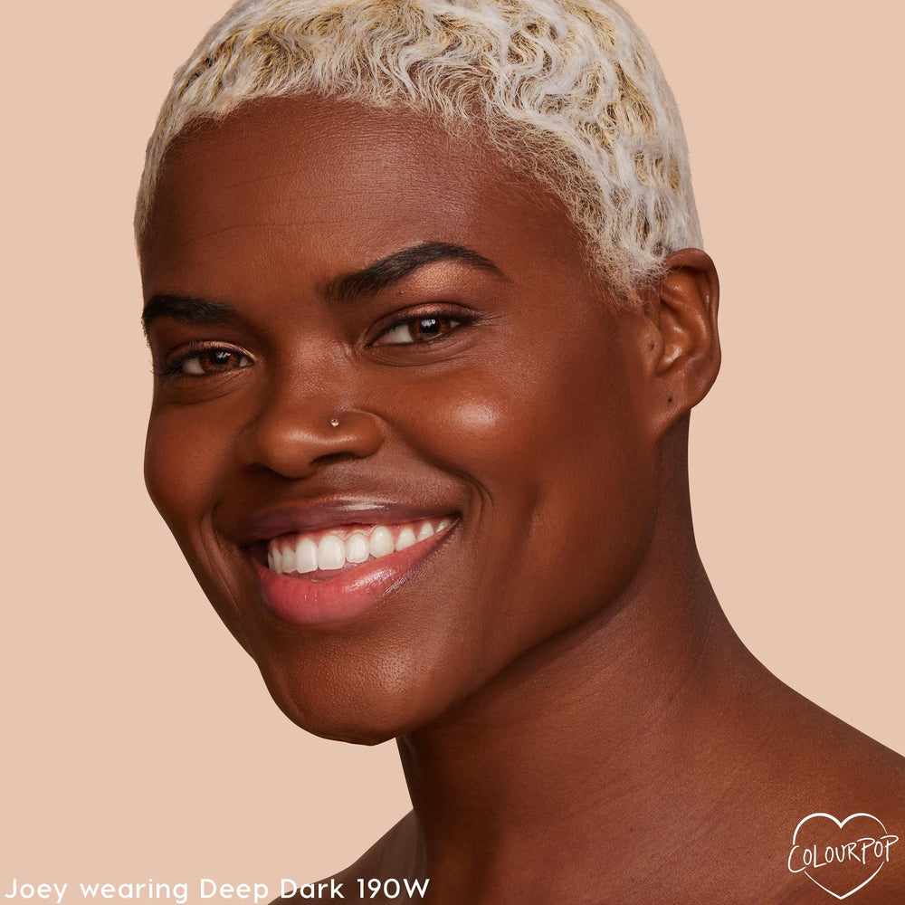 Model wearing ColourPop Pretty Fresh Creamy Concealer in Deep Dark 190 W smiles, showcasing smooth, radiant coverage in a close-up portrait against a warm beige studio background.