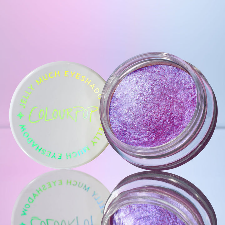 Open jar of ColourPop Jelly Much Shadow, Duckling, revealing metallic lavender‑pink shimmer in a clear pot, holographic ColourPop lid beside it, styled on a reflective surface with a pastel blue‑pink gradient backdrop.