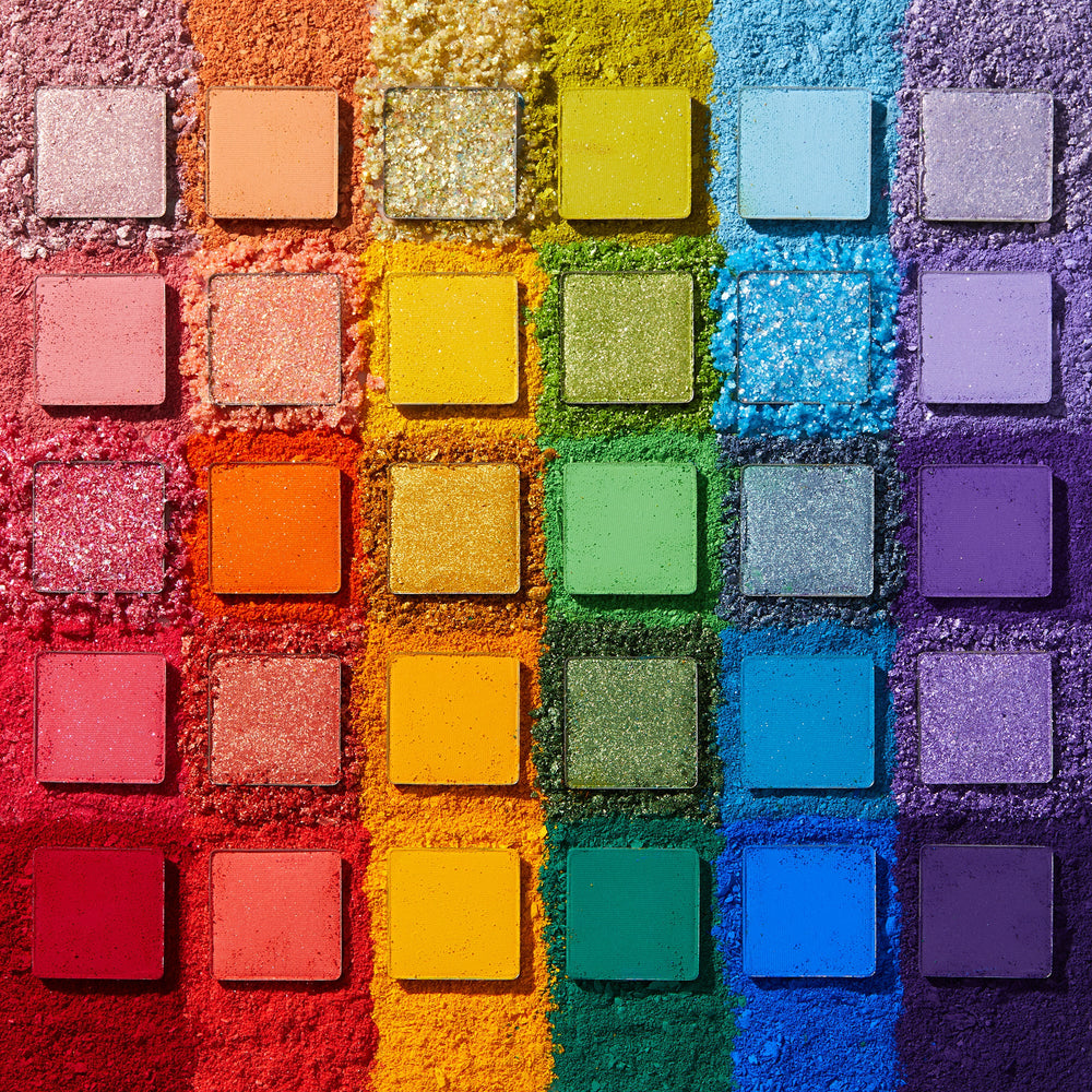 Fade Into Hue shadow palette displays 30 square pans in a rainbow grid - matte, shimmer, and pressed glitter - set on crushed pigment in a deconstructed flat lay; a vibrant ColourPop Shadow Palette.