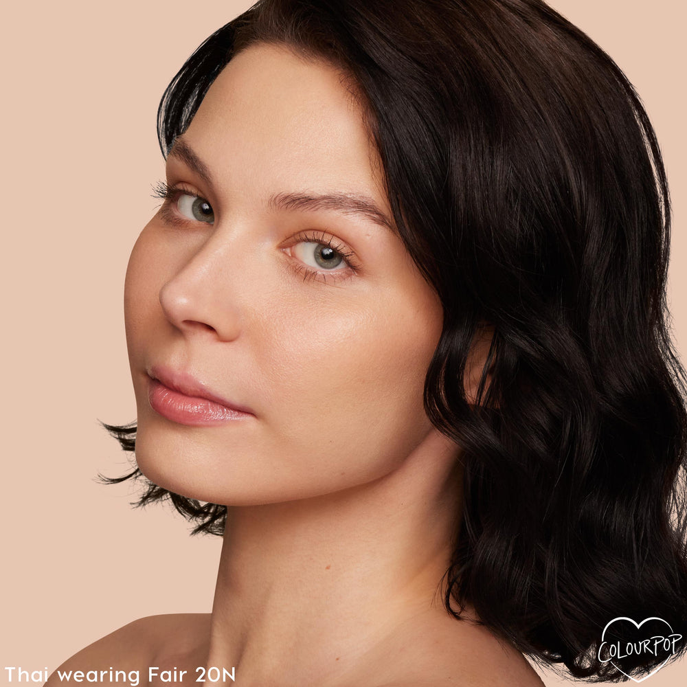 Model with short dark wavy hair and natural, radiant skin wearing Fair 20 N in ColourPop Pretty Fresh Creamy Concealer, tilting head in a studio close-up on a peach background, showcasing smooth under-eyes and seamless coverage.
