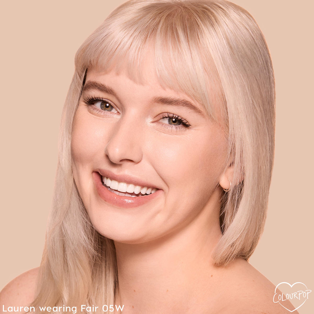 Model wearing Fair 05 W shade of ColourPop Pretty Fresh Creamy Concealer smiles in a close-up, showing luminous, even skin and bright under-eyes against a neutral beige background, highlighting natural, hydrating coverage.