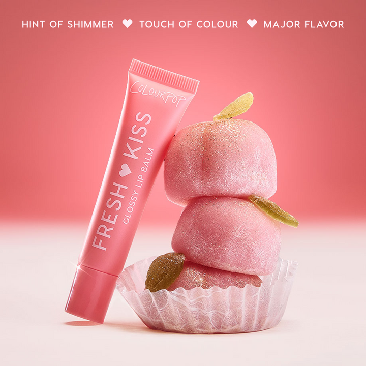 Colourpop Fresh Kiss Glossy Balm in Peach Mochi leaning against stacked pink mochi candies with sugar crystals, highlighting the shimmery Lip Gloss & Lip Oil hybrid on a soft red and white background.