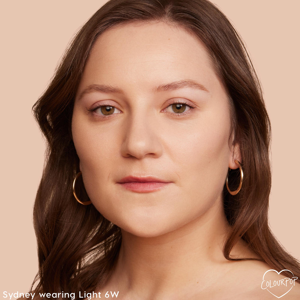 Model wearing ColourPop Pretty Fresh Tinted Moisturizer in Light 6 W, showing smooth, natural coverage on a close-up face with gold hoop earrings against a peach background.