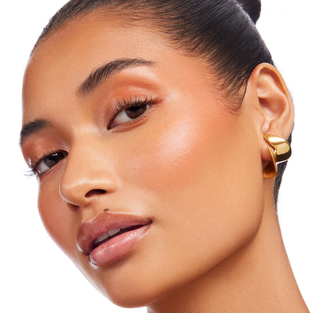 Ignite Lite Stix cream highlighter from ColourPop Lite Stix sweeping a peachy-gold glow across a model’s cheekbones and brow bone, catching light in a close-up with glossy lips and a gold earring against a clean white background.