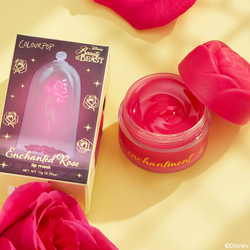 Enchanted Rose ColourPop Lip Mask jar opened to reveal glossy pink balm, rose-shaped lid set aside, beside Disney Beauty and the Beast box with enchanted rose bell graphic, styled on a yellow surface with red rose petals.