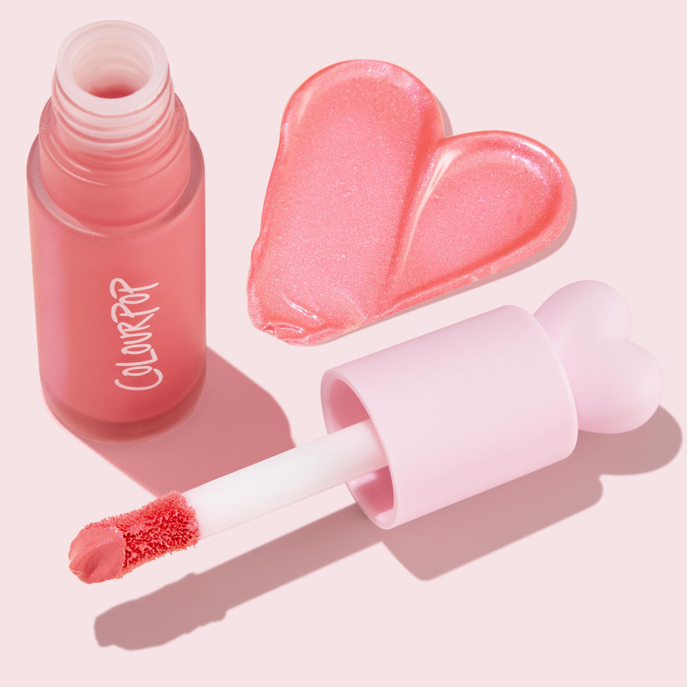 ColourPop Liquid Blush I Love It swatches a shimmery pink heart, with the doe-foot applicator and heart-topped pastel cap next to a frosted rosy bottle on a soft pink background.