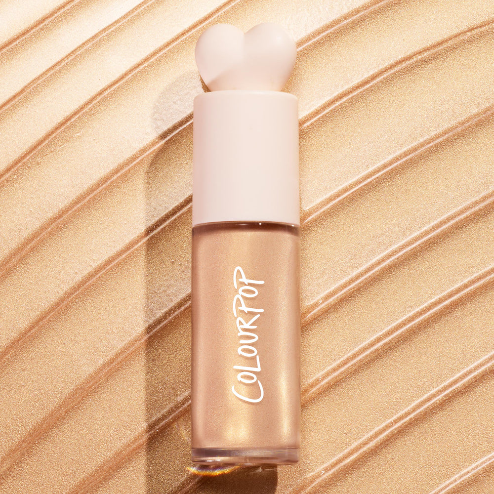 ColourPop Liquid Highlighter Immediately Yes stands upright with a heart-shaped ivory cap and frosted tube, showing a champagne-gold sheen against sweeping metallic swatches, spotlighting the product’s radiant, blendable glow.