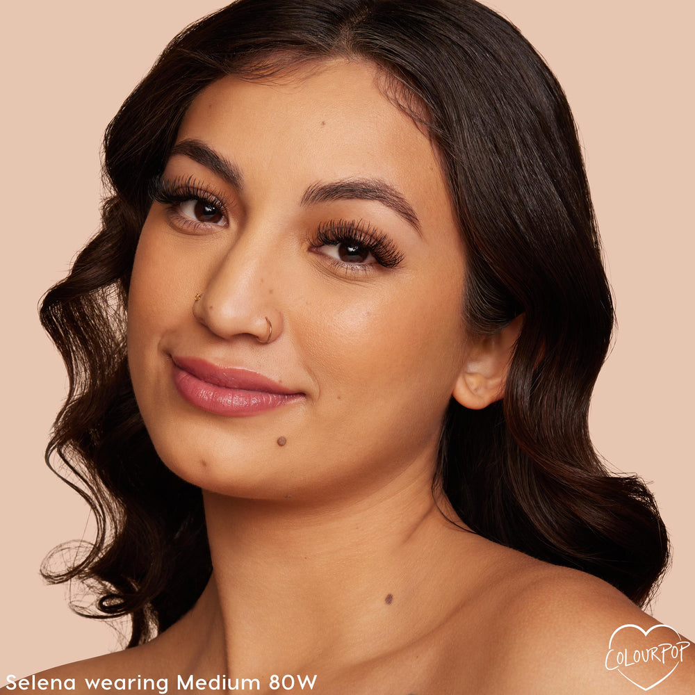 Model wearing ColourPop Hyaluronic Hydrating Foundation in Medium 80W, a warm medium shade delivering dewy, even coverage; close-up portrait shows smooth complexion, full lashes, and neutral lips against a beige studio background.