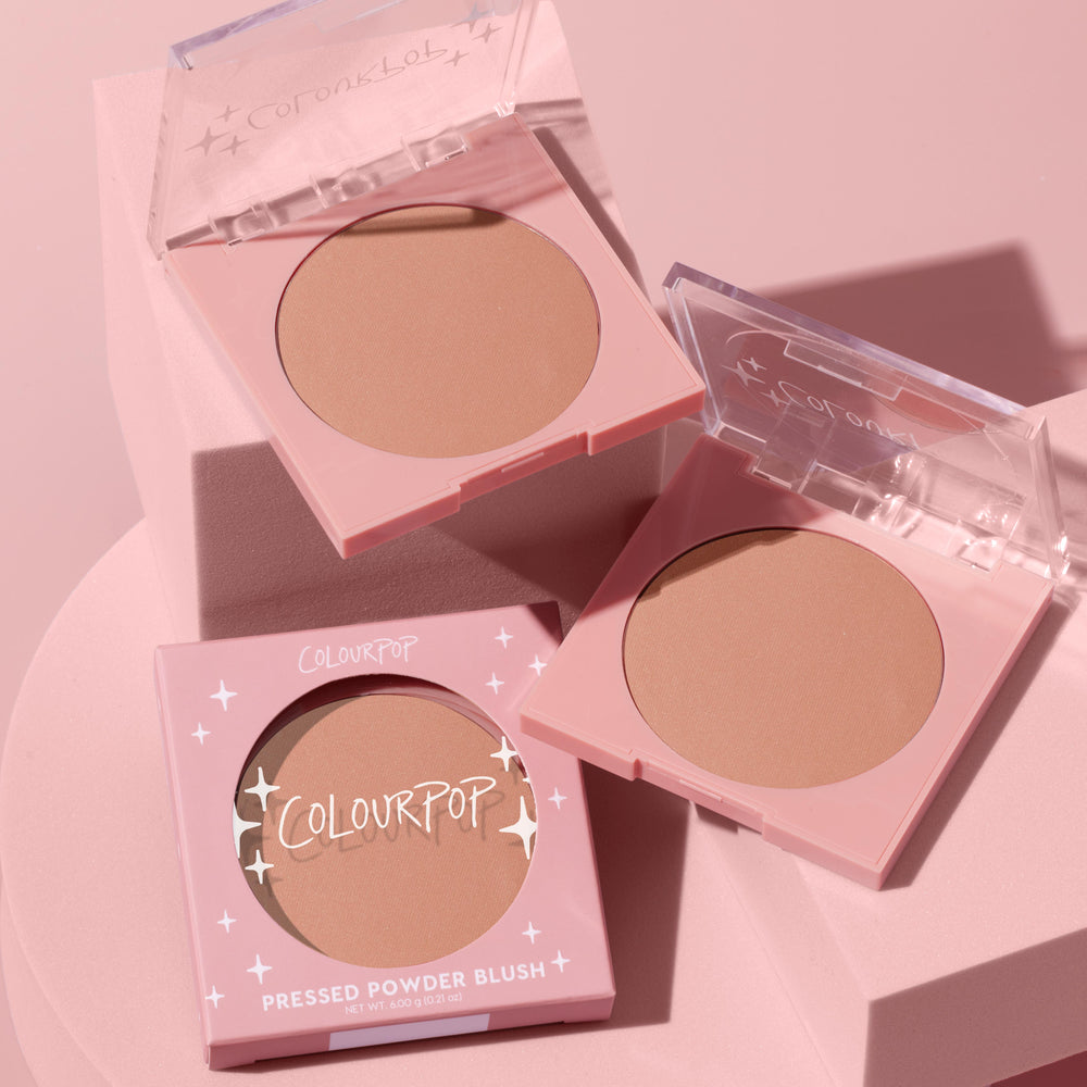 ColourPop Pressed Powder Blush in Out & About displayed in pink compacts with clear flip lids, warm peach‑nude matte pans, and one boxed unit, arranged on soft pink blocks in a clean product flat lay.