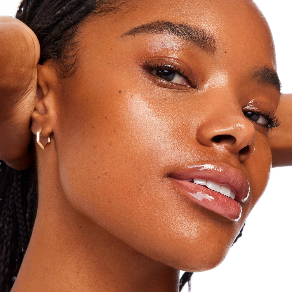 Model tilts face in a bright studio, skin dewy and luminous to showcase Selfie Ready Serum Stick by ColourPop Serum Stick, with glossy lips and a small gold geometric hoop earring against a clean white background.