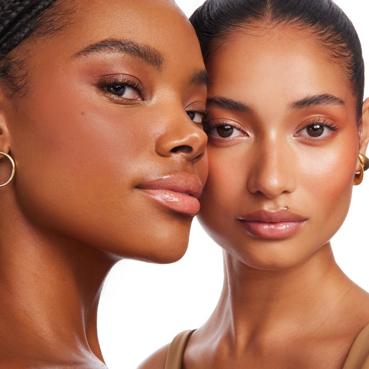 Two models showcase a warm, sun-kissed glow as ColourPop Bronzer Stix in Rincon Point blends across cheeks and temples, delivering a natural bronze sheen on diverse skin tones in a studio close-up with glossy lips and gold hoop earrings.