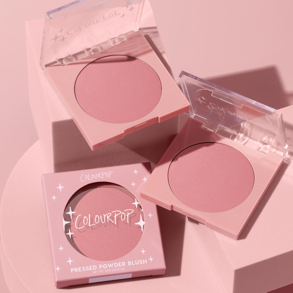 ColourPop Pressed Powder Blush in Sassy Pants, a matte dusty-rose hue, displayed in two open pastel-pink square compacts with clear lids beside the boxed unit with star accents, styled on a monochromatic blush-pink pedestal.
