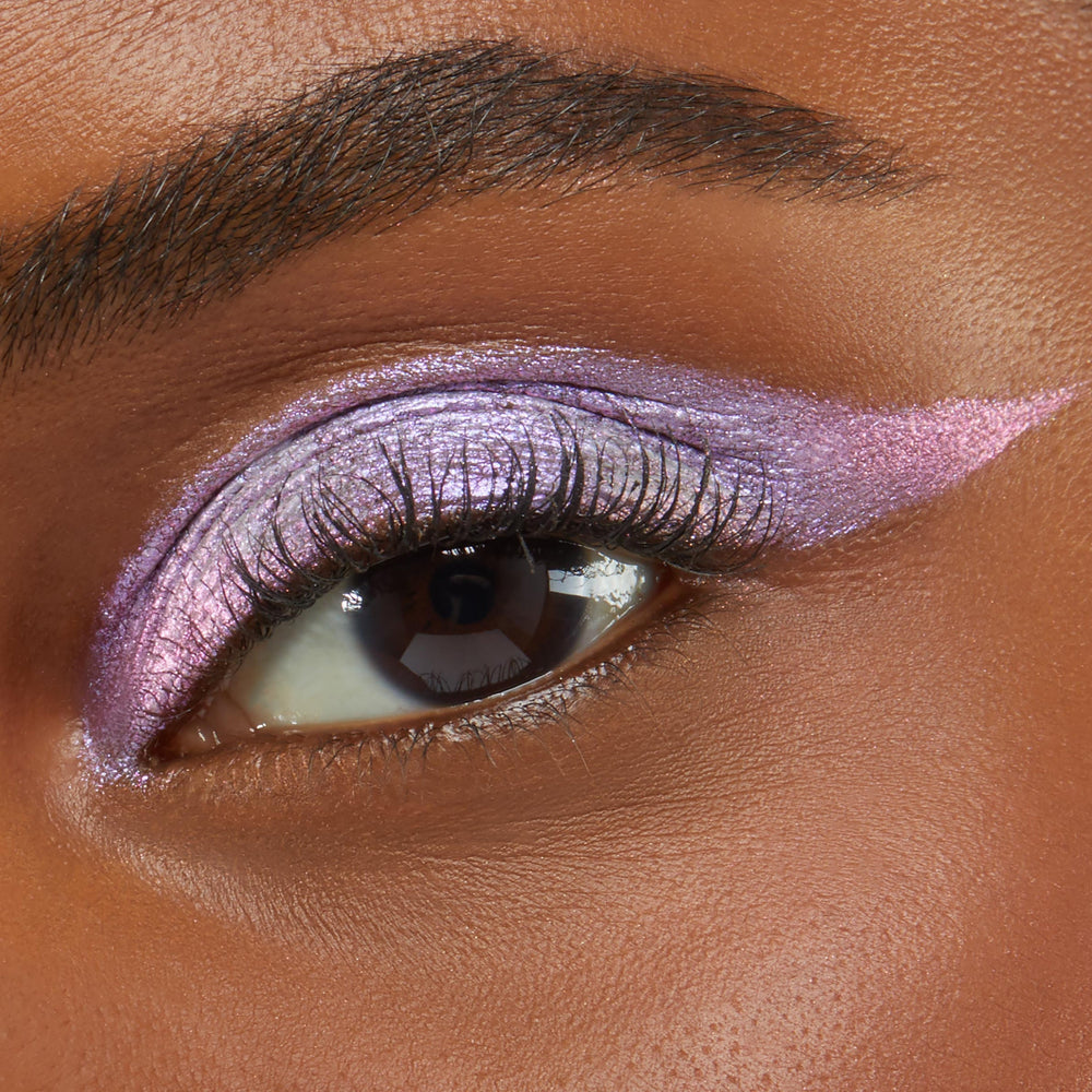 Close-up of an eye wearing Colourpop Cotton Candy Shadow Stix, showing shimmering pastel pink and lavender cream eyeshadow blended across the lid and extended into a sharp wing on smooth medium-brown skin.