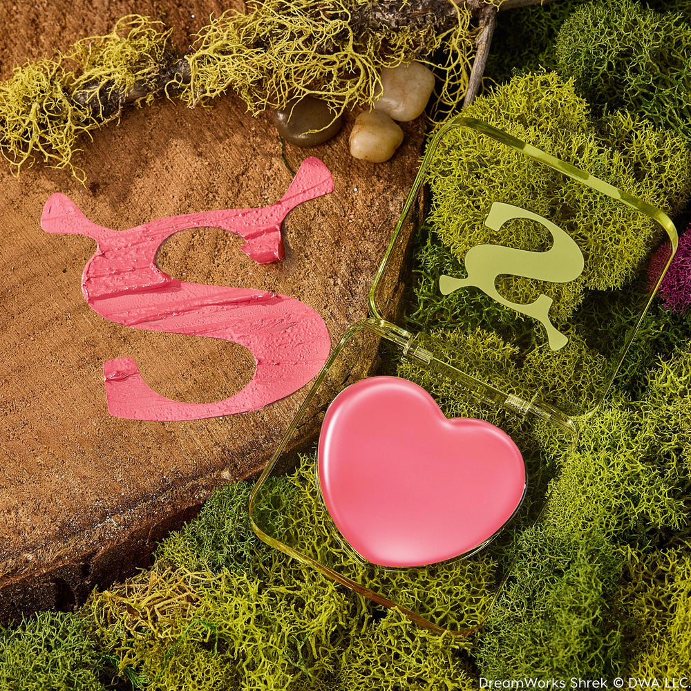 Heart-shaped pink balm in a translucent green compact sits open on moss and a wood slab with a Shrek-style S, showcasing ColourPop Lip and Cheek Balm, True Love’s Form, a lip and cheek tint in a playful swamp-inspired setting.