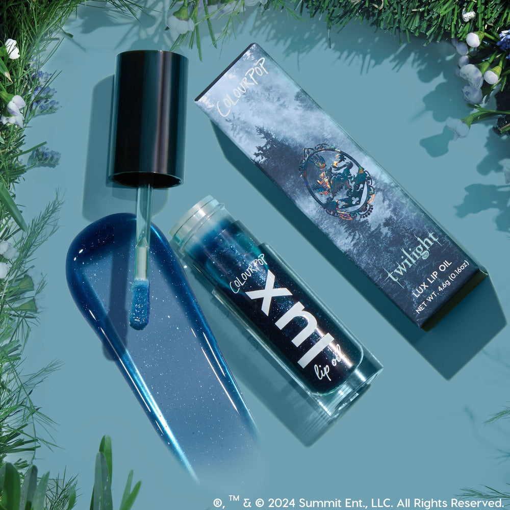 Team Edward Lux Lip Oil swatched: a sparkling deep blue, shimmer-packed gloss. Doe-foot applicator, open ColourPop Lux Lip Oil tube, and Twilight collab box rest on a teal surface framed by evergreen sprigs.