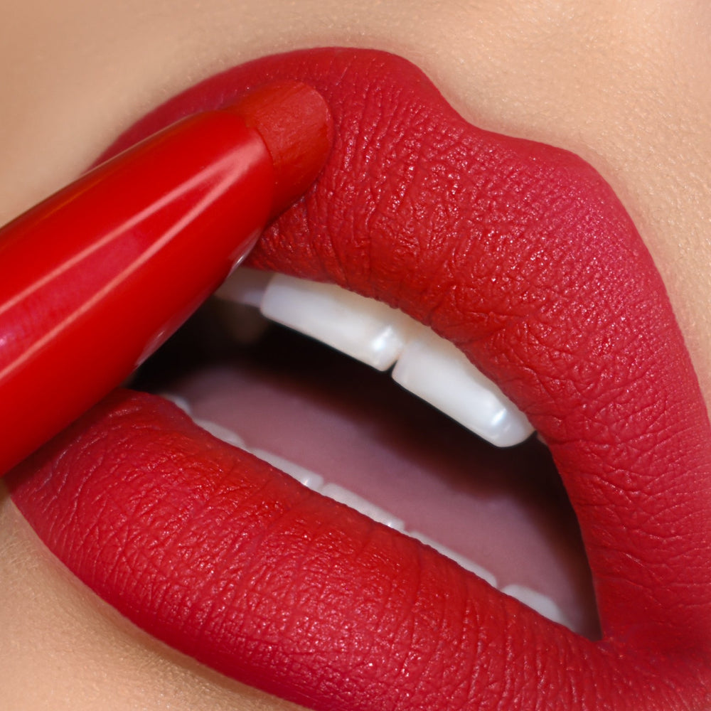 ColourPop Lippie Stix in Trust Me glides onto full lips, laying down vivid true-red matte color; extreme close-up shows the bullet tracing the upper lip with crisp edges, smooth texture, and white teeth peeking for a bold finish.