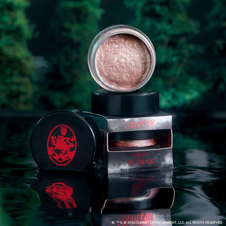 ColourPop Cullen Coven Jelly Much Shadow, a shimmery rose-bronze cream eyeshadow from the Twilight collection, opened and stacked on its Eclipse packaging against a dark, misty forest backdrop with reflections on glossy black water.