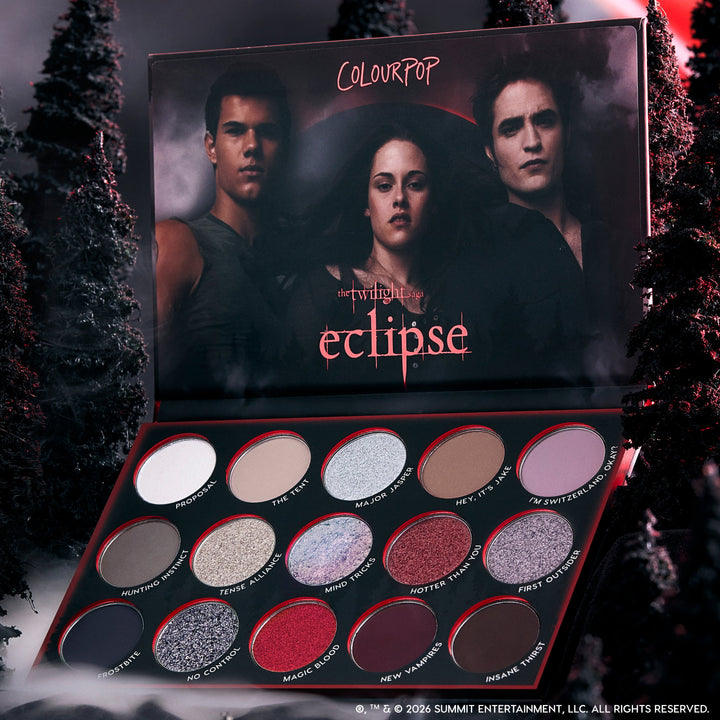 ColourPop Eclipse shadow palette from the Twilight collection lying open in a dark forest setting, showing 12 shimmer and matte cool-toned and red eyeshadows arranged in rows beneath moody character artwork on the inner lid.
