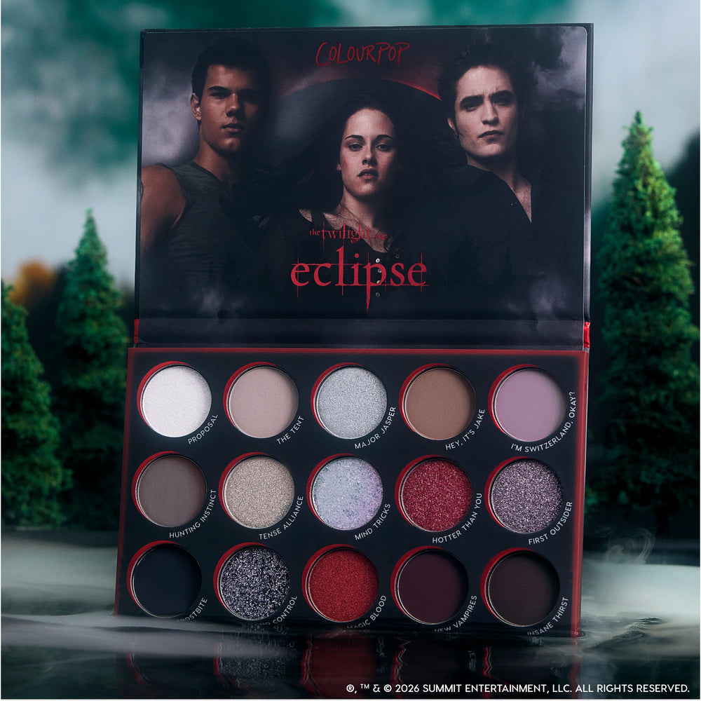 ColourPop Eclipse shadow palette from the Twilight collection standing open by a misty forest, showing 15 vampy and neutral shimmer and matte eyeshadows with red-trimmed pans and moody character artwork on the lid.