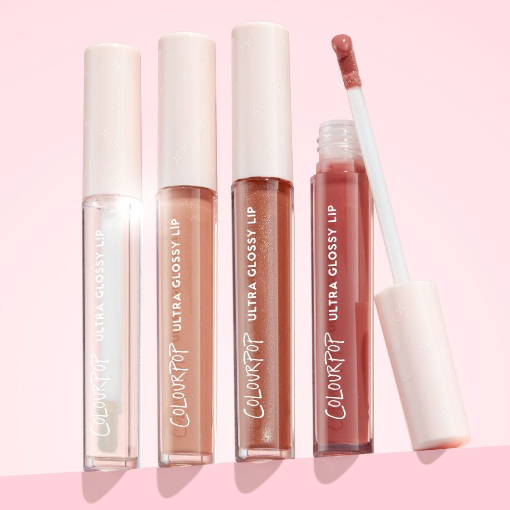 ColourPop Ultra Glossy Lip 20 Carats with doe‑foot applicator lifted, showing rosy nude shine, stands beside three closed ColourPop Ultra Glossy Lip tubes - clear, peach nude, and bronze shimmer - with star‑embossed caps on a soft pink backdrop.