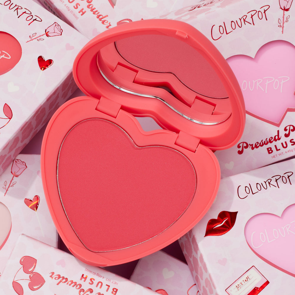 Heart-shaped Woo Me compact opens to reveal a rosy-coral pan and mirror, showcasing ColourPop Pressed Powder Blush in Valentine-themed packaging with hearts and lips, surrounded by stacked pink boxes for a cute gift-ready look.