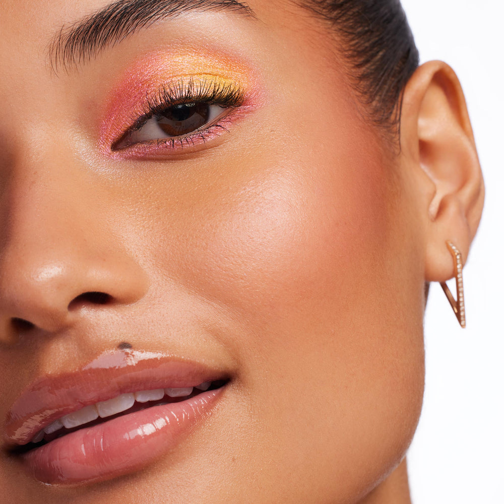 ColourPop Pressed Powder Blush Single in New to U adds a soft peachy glow on a model’s cheek, close-up studio shot with glossy nude lips and pink-to-gold shimmer eyeshadow, showcasing the blendable payoff and smooth finish.
