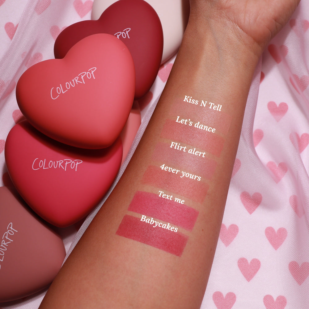 Swatched forearm displays “Text Me” ColourPop Pressed Powder Blush, a rosy pink matte stripe, alongside shades Babycakes, 4ever Yours, Flirt Alert, Let’s Dance, and Kiss N Tell; heart-shaped ColourPop compacts on pink heart-pattern fabric.