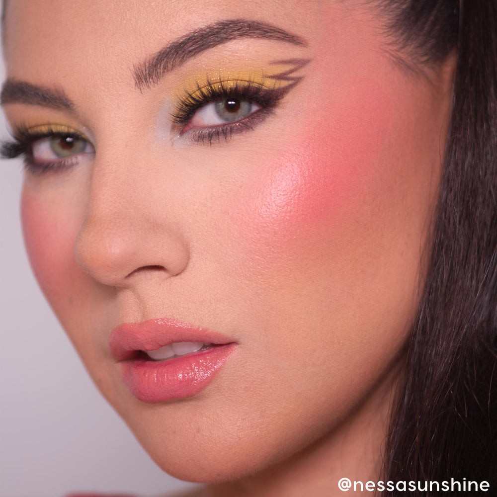 Electrifying Dreams ColourPop Cream Blush delivering a dewy coral-pink glow on high cheekbones, shown in a close-up face with bold yellow graphic winged eyeliner, long lashes, and glossy pink lips under soft studio lighting.